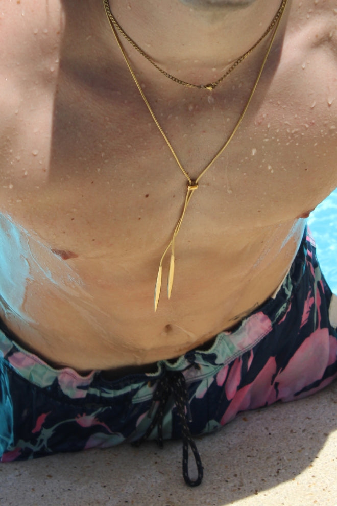 Collar Viento Tico visto en primer plano sobre el pecho de un hombre con bañador colorido, mostrando la cadena dorada y los colgantes alargados en un entorno de piscina.